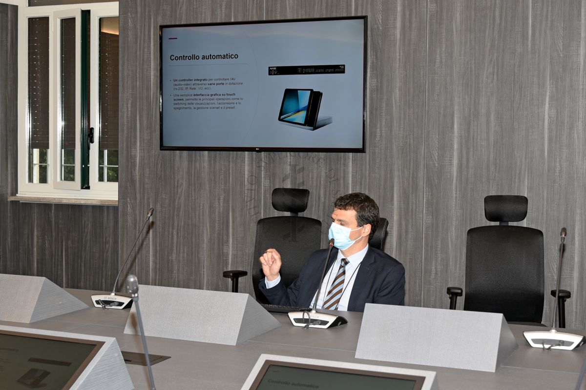 Il Dr. Corrado Di Benedetto, seduto al tavolo nella Sala Crisi dell'ISS, mentre spiega le funzionalità multimediali della sala, visibili sullo schermo dietro di lui