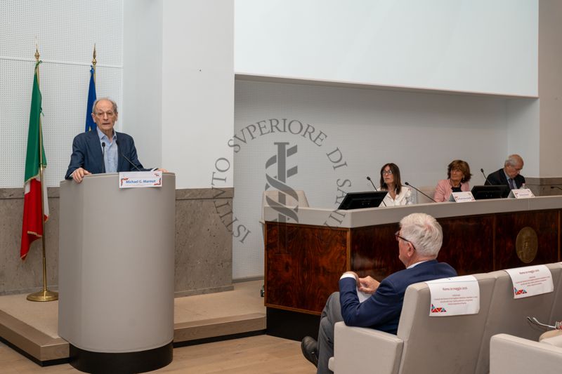 Intervento in Aula Magna dell'ISS di Sir Michael Gideon Marmot, professore di epidemiologia e sanità pubblica presso l'University College di Londra e direttore dell'UCL Intitute of Health Equity; alla sua sinistra si possono notare i moderatori: Dott.ssa Raffaella Bucciardini, direttore di reparto del Centro Nazionale per la Salute Globale dell'ISS; Dott.ssa Serena Donati direttore del reparto della Salute della Donna e dell'Età Evolutiva (CNaPPS dell'ISS); e il Dr. Leonardo Speri, noto psicologo, psicoterapeuta e psicosocioanalista italiano, attualmente presidente dell'associazione Ariele Psicoterapia e docente presso la Scuola di Specializzazione in Psicoterapia della COIRAG (Confederazione delle Organizzazioni Italiane per la Ricerca Analitica sui Gruppi).