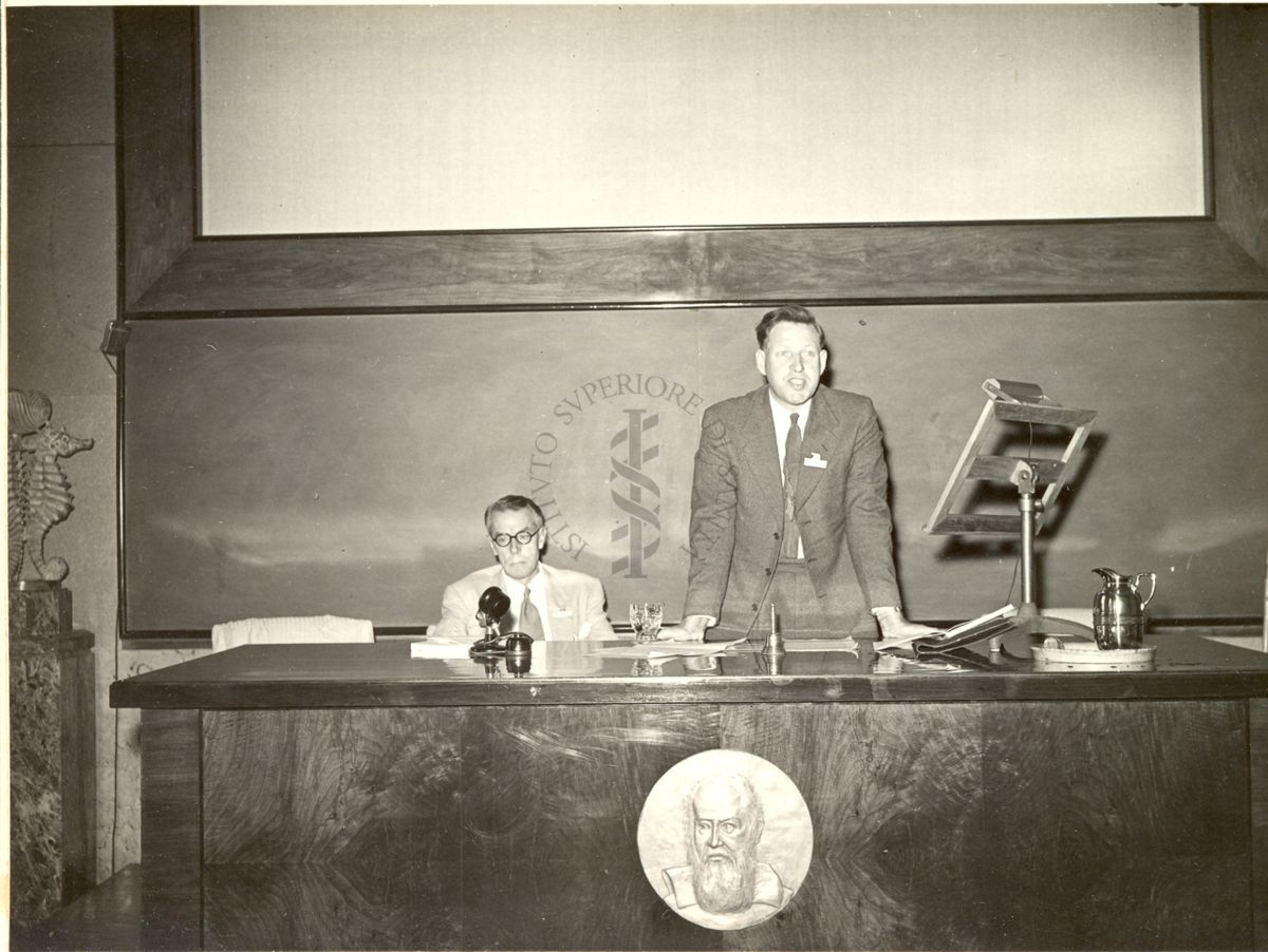 Il Prof. Frederick Charles Bawden ripreso in piedi dietro un leggio posto su un tavolo, durante la relazione tenuta al VI Congresso Internazionale di Microbiologia. Alla sua destra, seduto al tavolo, il Prof. K. N. Smith.