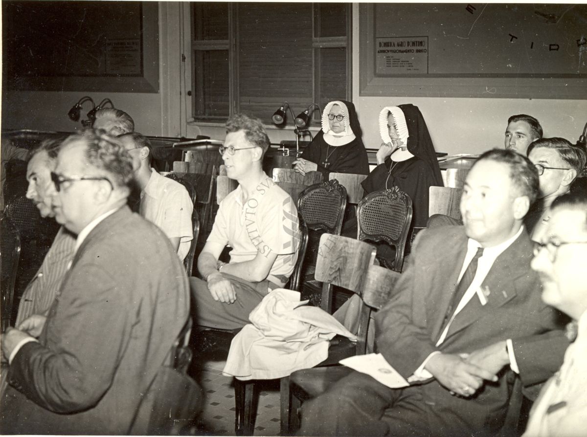 Ripresa di parte dell'uditorio seduto su sedie durante le relazioni al VI Congresso Internazionale di Microbiologia. Inquadrati: il Prof. Norman Wingate Pirie (terzo da destra in seconda fila) e Suor Marie Susanne (ultima a sinistra in ultima fila)