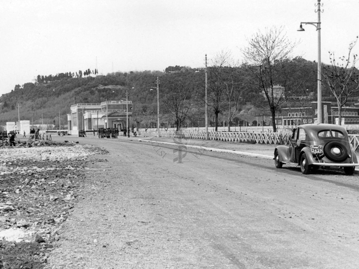 Inquadratura di una carreggiata che corre parallela al Tevere, con in fondo il Palazzo Littorio in costruzione, sulla quale transita un'autovettura targata Roma (numeri targa non visibili)