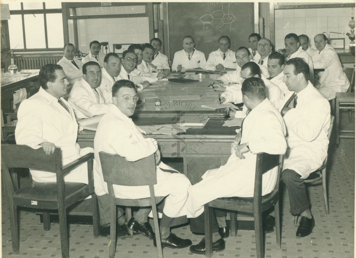 Foto di gruppo dei partecipanti al corso sugli stupefacenti, in camice, seduti intorno a un tavolo all'interno di un'aula dell'Istituto Superiore di Sanità, sullo sfondo una lavagna e alcuni strumenti da laboratorio