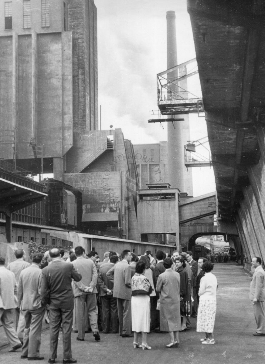 Un gruppo di uomini e donne ripresi di spalle all'esterno di una fabbrica. Sullo sfondo altri elementi architettonici industriali.
