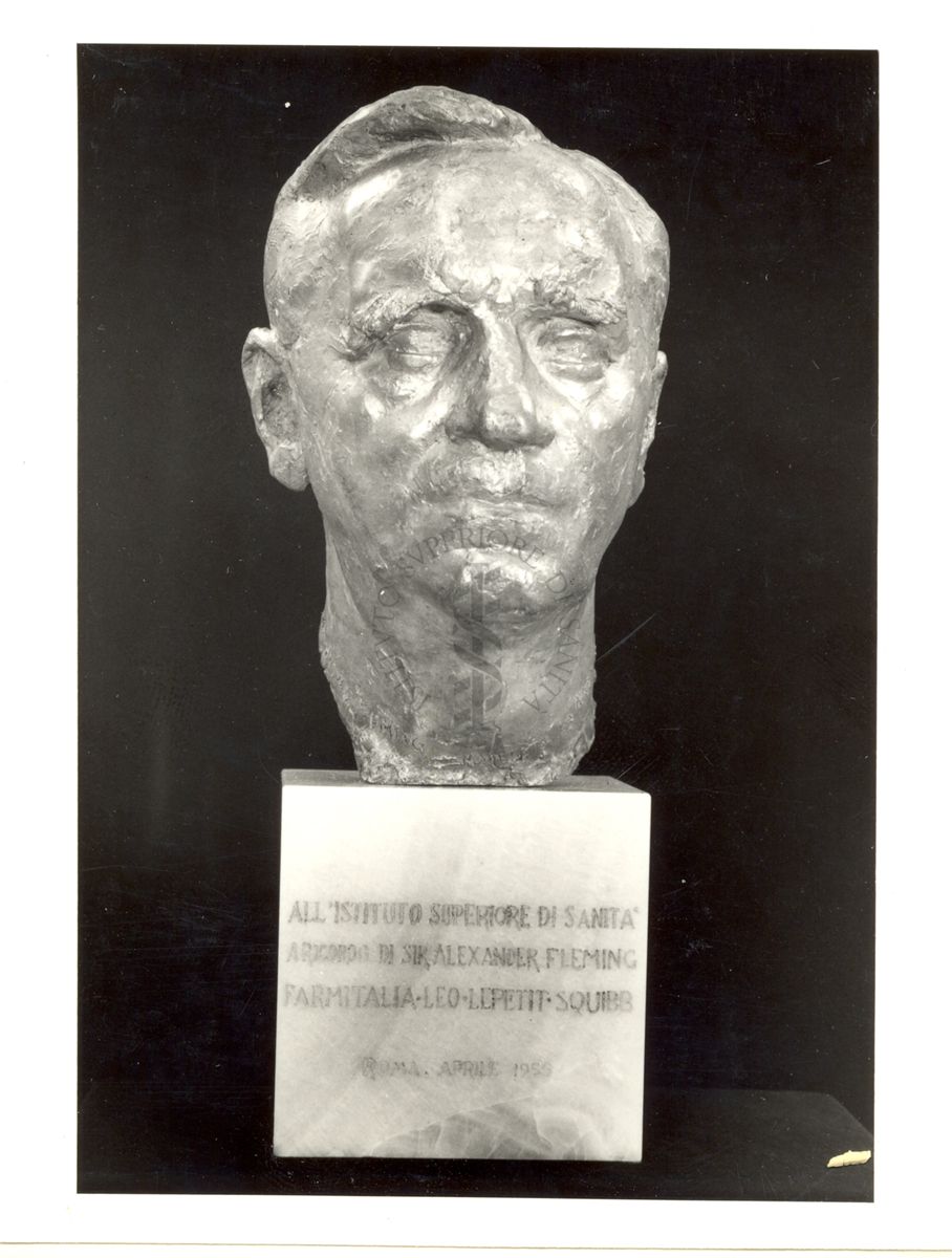 Fotografia, su fondo scuro, del busto di Sir Alexander Fleming, in bronzo dorato su base di marmo, donato all'Istituto Superiore di Sanità dalle aziende farmaceutiche Farmitalia, Leo, Lepetit e Squibb nel aprile 1955