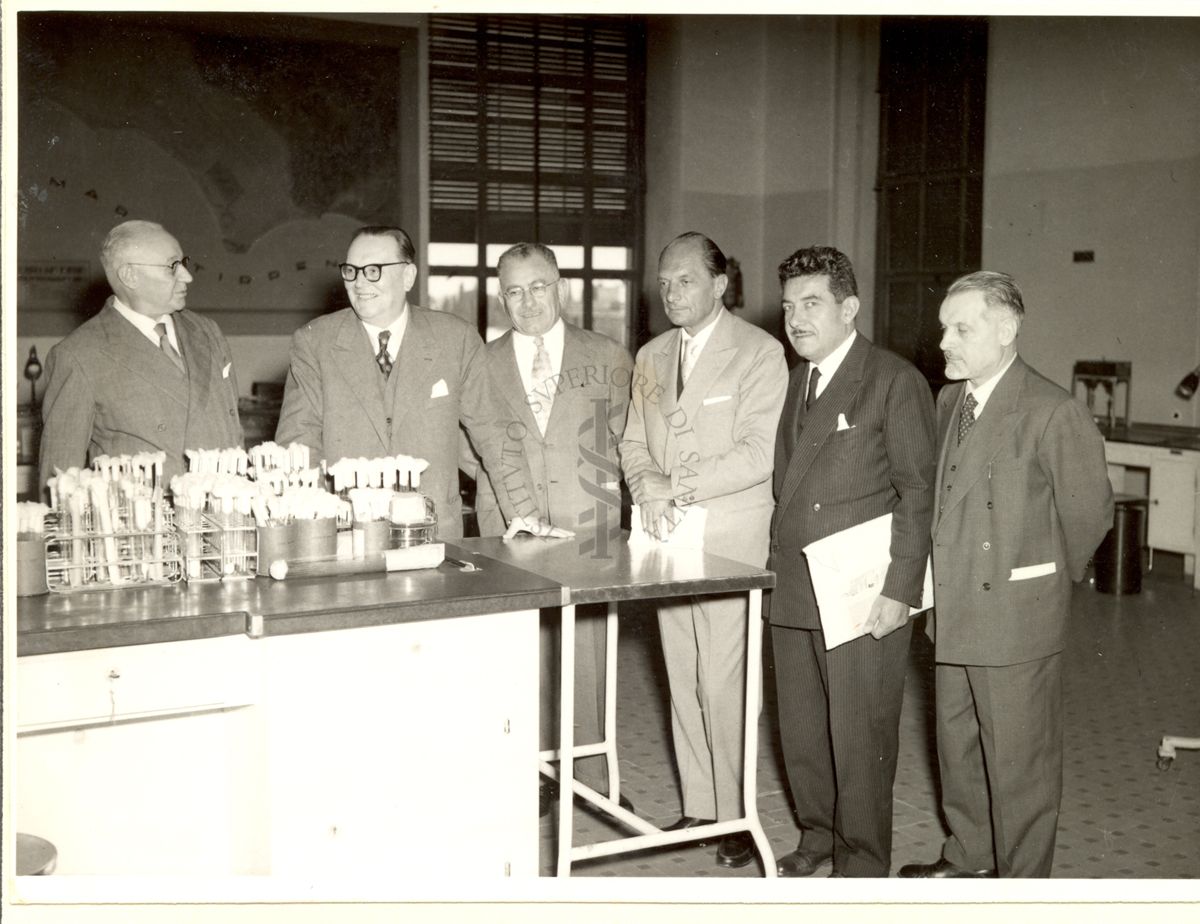 Foto di gruppo al banco di un laboratorio dell'Istituto Superiore di Sanità.
Da sinistra: il Prof. Domenico Marotta, il terzo On. Luigi Carmagnola, l'On. Piero Montagnani Marelli, l'On. Giuseppe Roda e l'On. Giuseppe Alberti