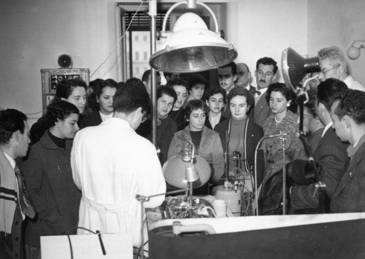 Il gruppo degli studenti uruguaiani in visita all'Istituto Superiore di Sanità all'interno di uno dei laboratori, attorno ad un  banconi di lavoro. Presenti due dipendenti dell'Istituto in camice bianco.