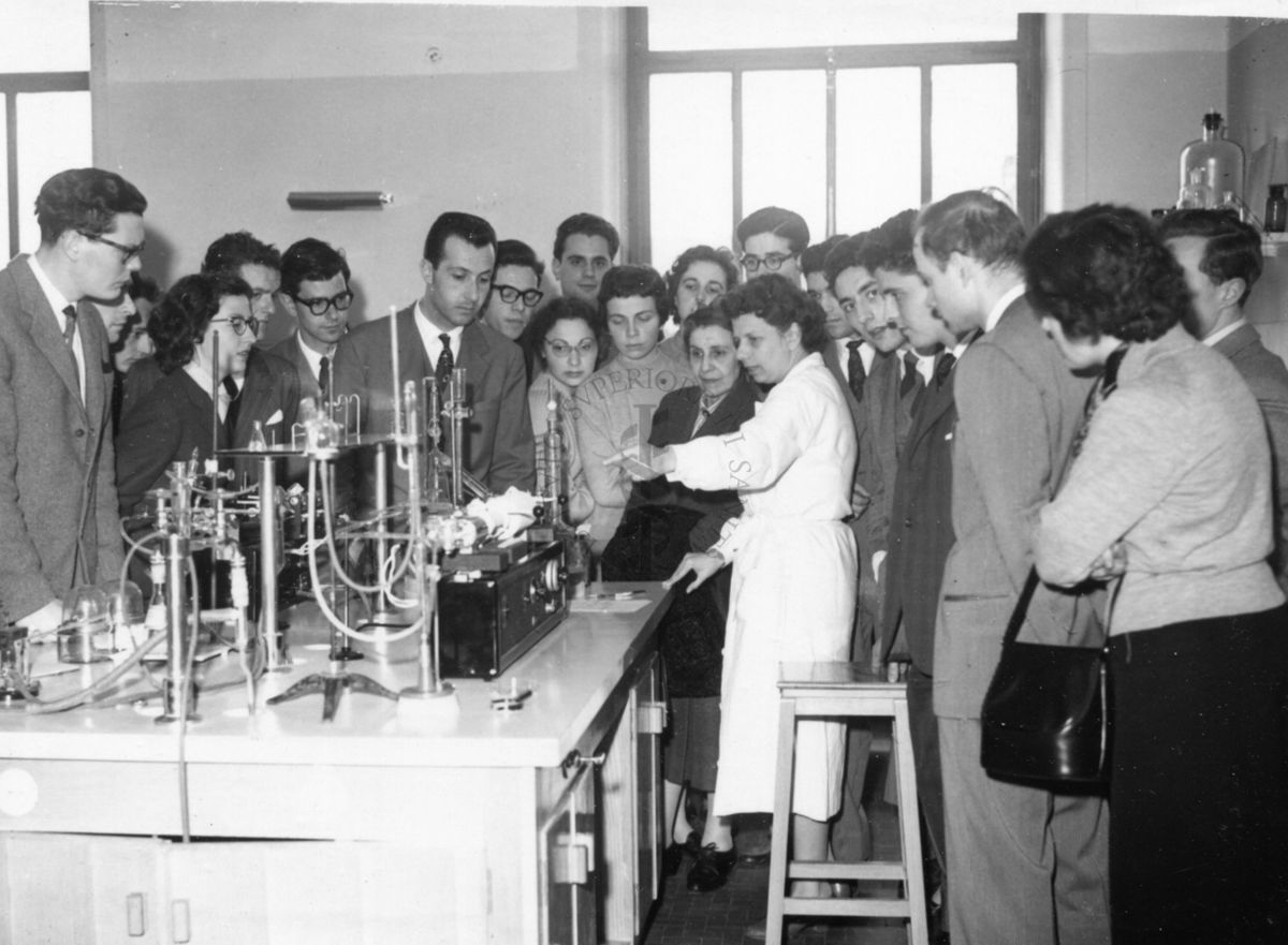 Il gruppo degli studenti della facoltà di medicina dell'Università di Firenze, in visita all'Istituto Superiore di Sanità, ripresi in uno dei laboratori attorno ad un bancone da lavoro e ad una dipendente dell'Istituto in camice bianco che illustra loro il funzionamento di un apparecchio