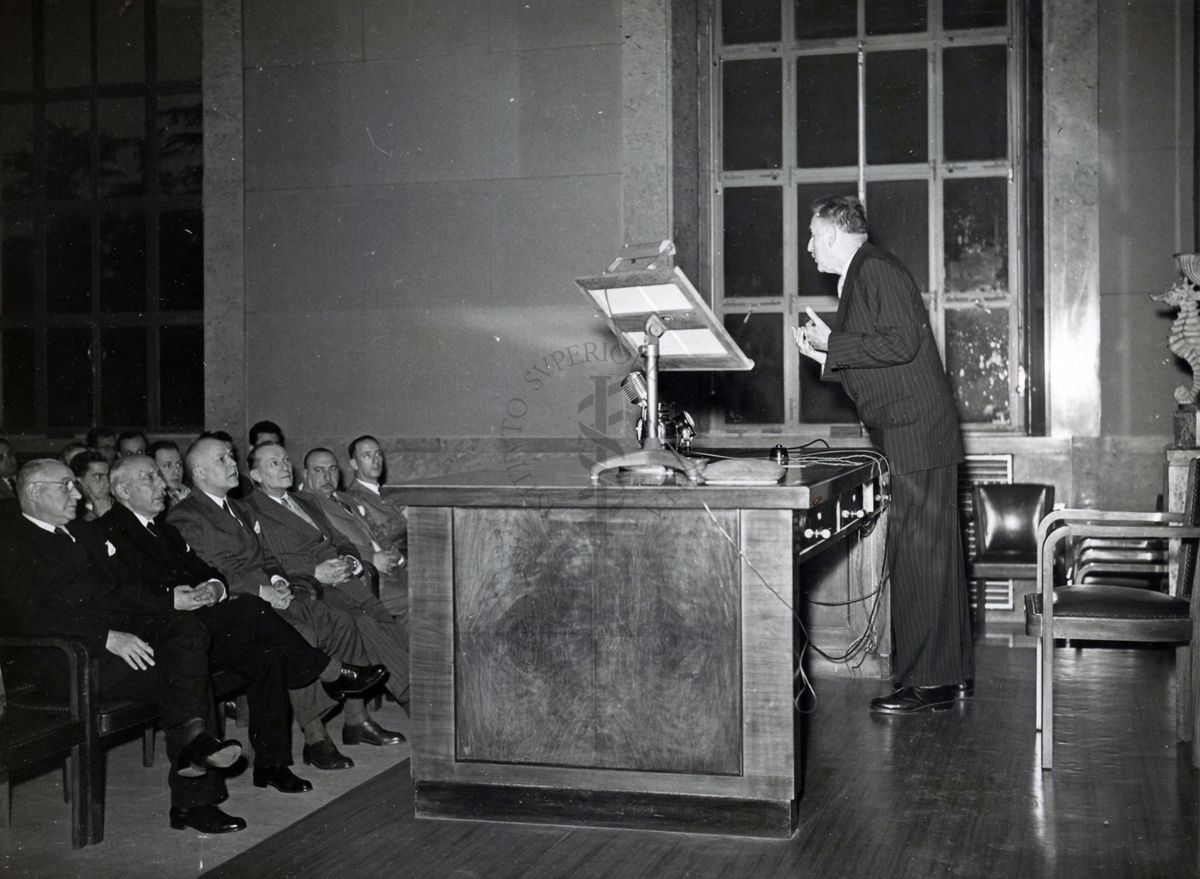 Il Prof. Antoine Lacassagne ripreso in piedi e di profilo, dietro al tavolo dei relatori, rivolto verso l'uditorio in occasione della sua conferenza nell'Aula magna dell'Istituto Superiore di Sanità