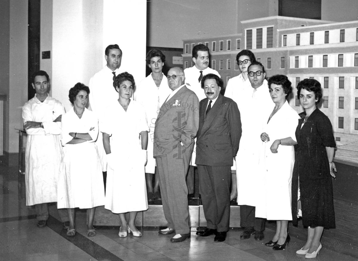 Foto di gruppo con al centro il Prof. Ernst Boris Chain e il Prof. Domenico Marotta e, primo da sinistra, in seconda fila, il Prof. Francesco Pocchiari. Sullo sfondo è visibile un plastico dell'edificio centrale di grandi dimensioni