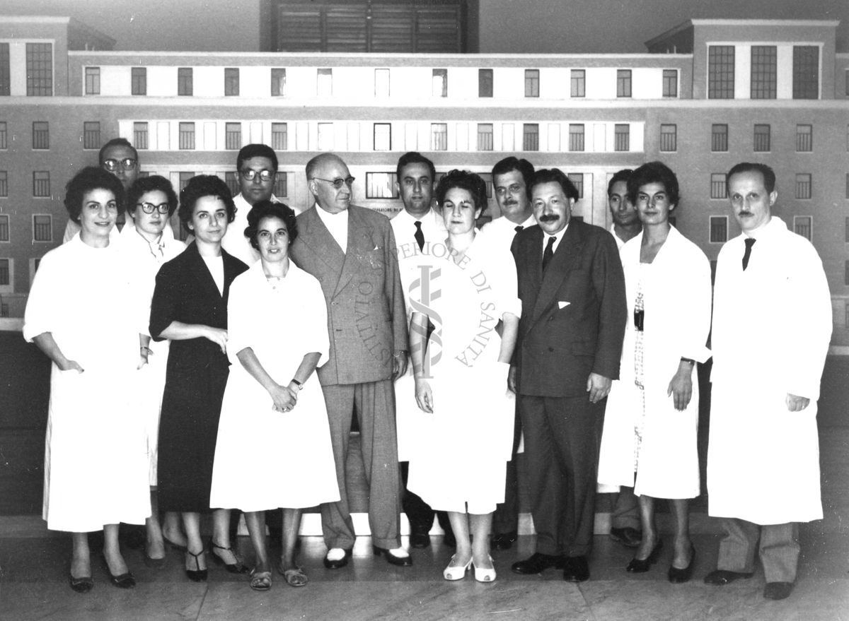 Foto di gruppo con al centro, senza camice, il Prof. Ernst Boris Chain e il Prof. Domenico Marotta e, in seconda fila, al centro e con il camice, il Prof. Francesco Pocchiari. Sullo sfondo è visibile un plastico dell'edificio centrale di grandi dimensioni.