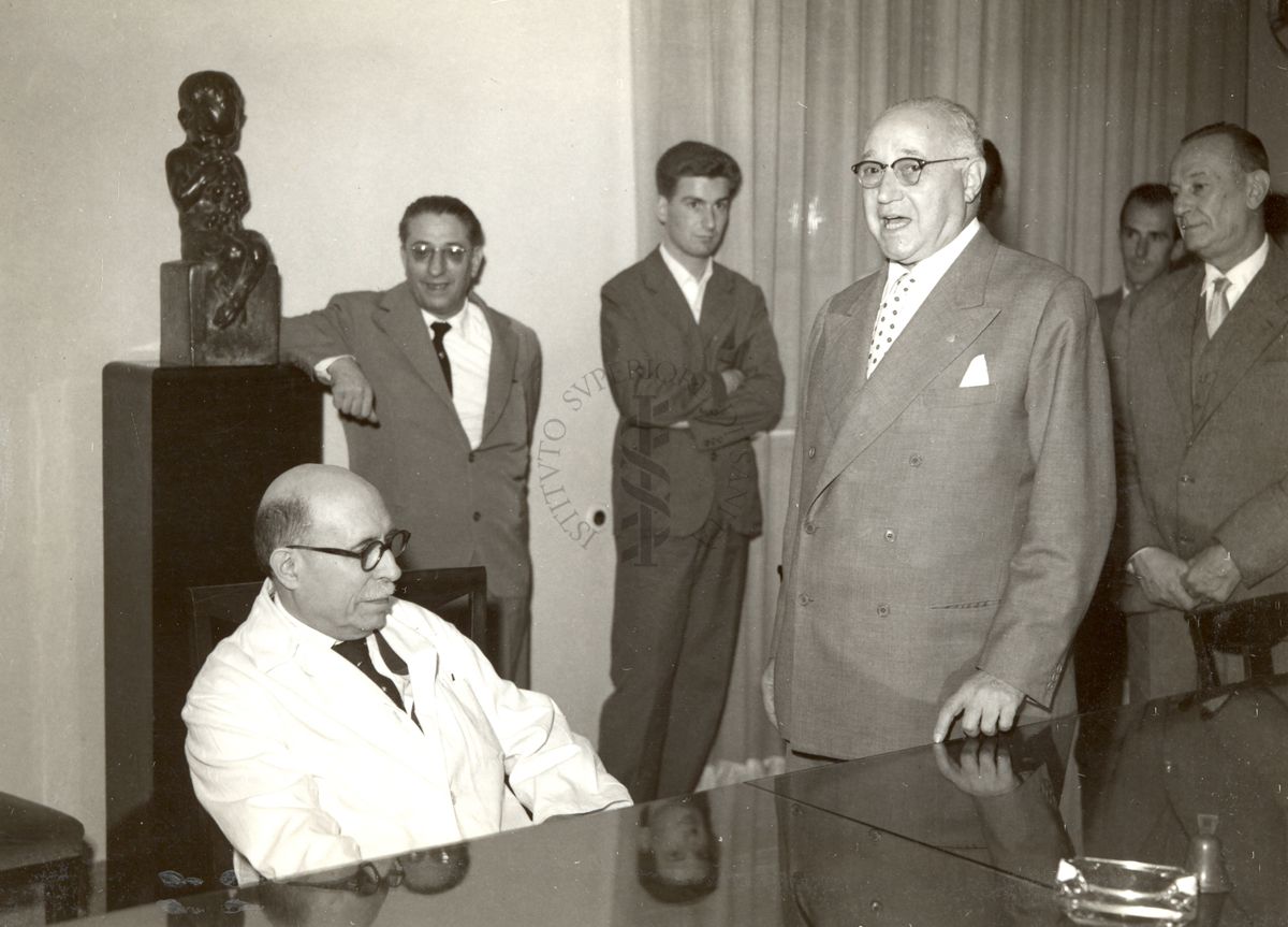 Il Prof. Domenico Marotta (in piedi a destra) annuncia la consegna della medaglia d'oro per i 50 anni di onorato servizio al Prof. Giulio Cesare Trabacchi (seduto con il camice); nella foto è presente anche il Prof. Massimo Pantaleoni (ultimo a destra)