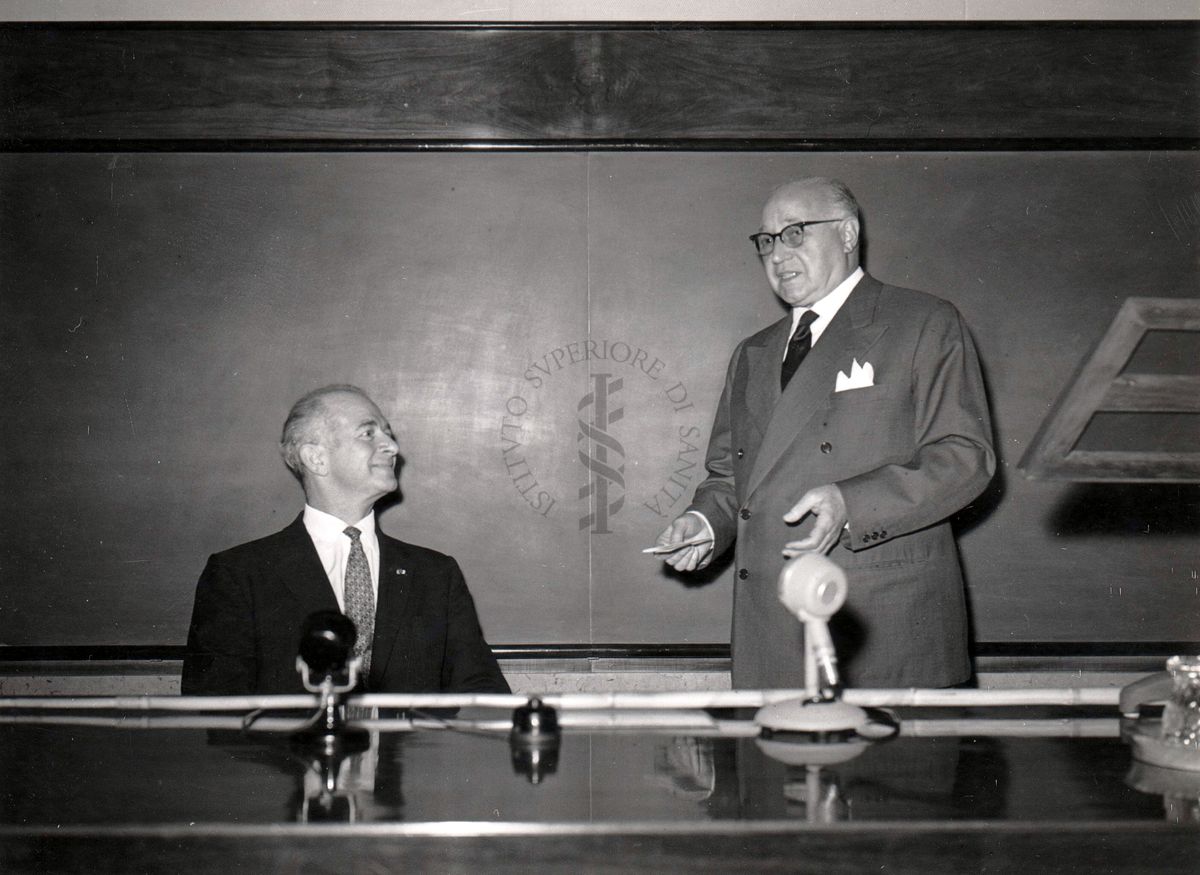 Il prof. Linus Carl Pauling (seduto, lato sinistro della foto) segue un discorso del prof. Domenico Marotta (in piedi, lato destro della foto) nell'aula magna dell'Istituto Superiore di Sanità.