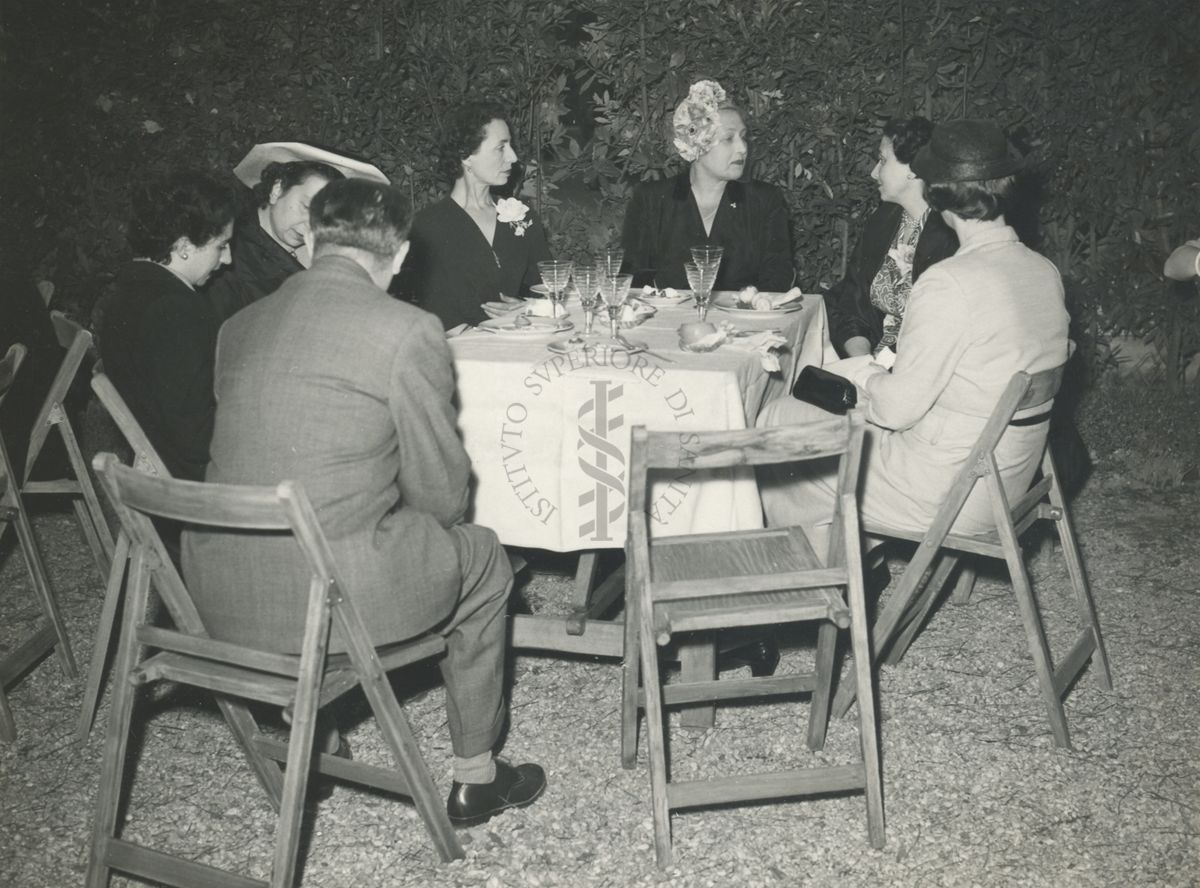Gruppo di sei donne ed un uomo (ripreso di spalle) seduti ad uno dei tavoli allestiti nel giardino dell'Istituto Superiore di sanità per il banchetto tenuto in occasione del IX Congresso internazionale dell'industria agraria