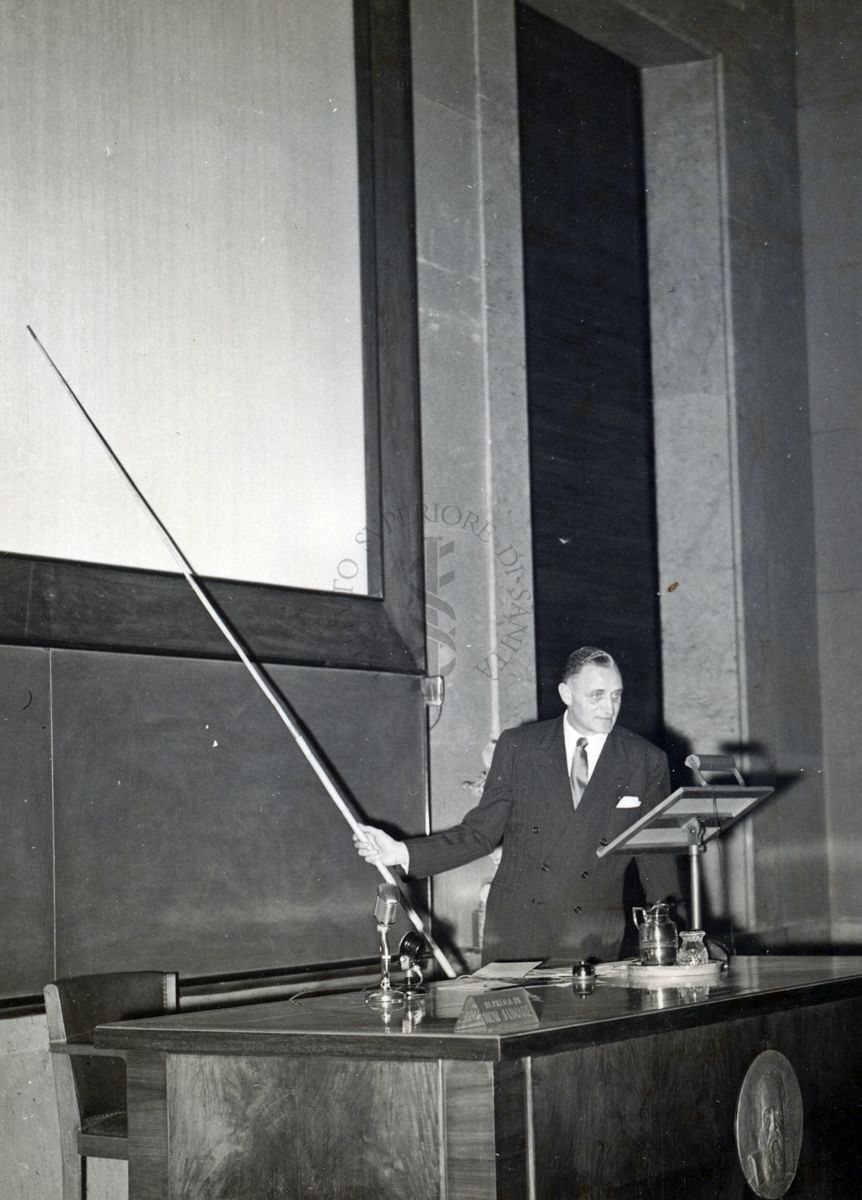 Il Prof. H. Corwin Hinshaw ripreso in piedi, dietro al tavolo dei relatori, mentre con un'asta indica la lavagna, durante la conferenza tenuta all'Istituto Superiore di Sanità.