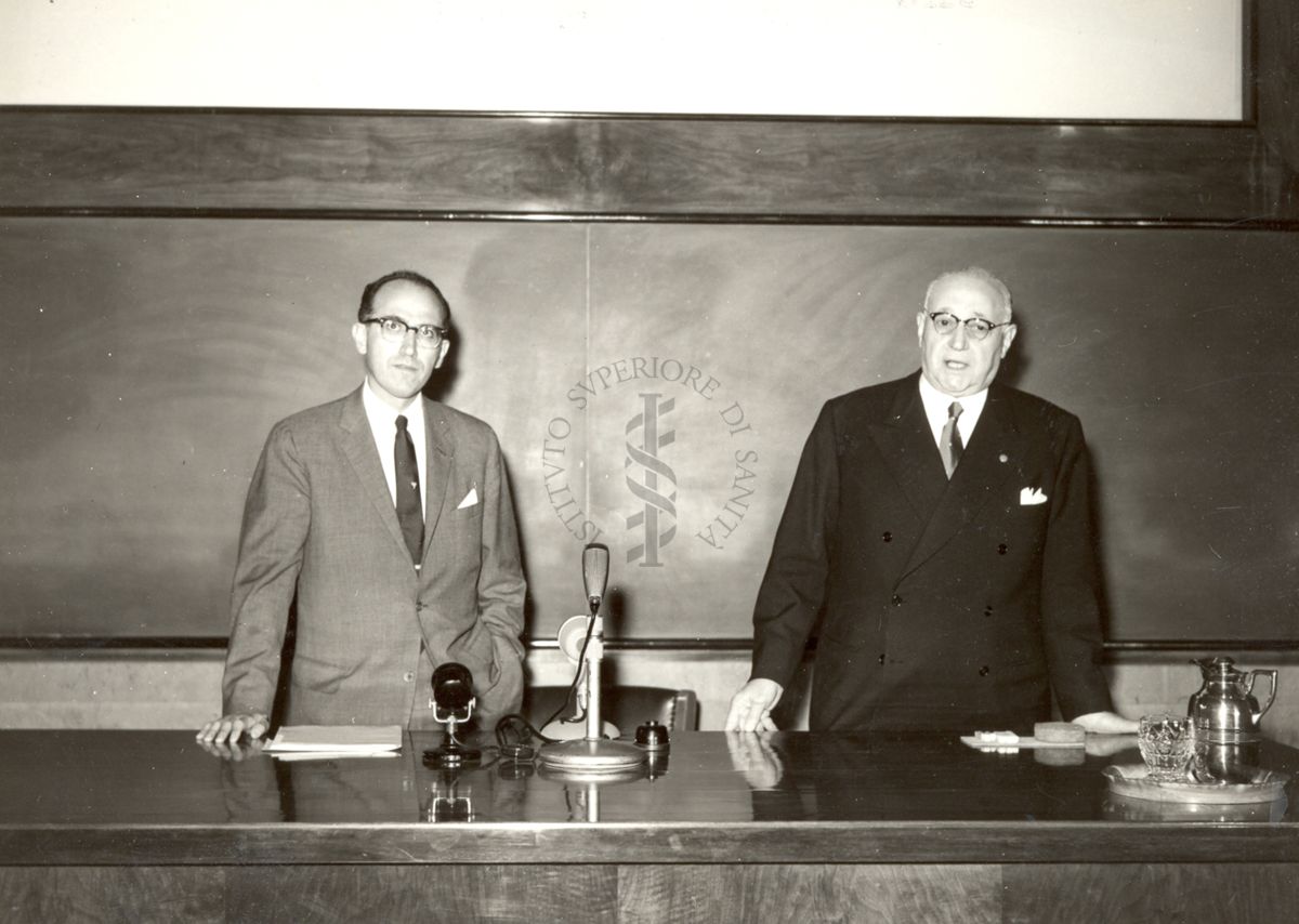 Il Prof. Salk (a sinistra) e il Prof. Marotta (a destra) in piedi dietro la cattedra dei relatori nell'aula magna dell'Istituto SUperiore di Sanità in occasione della conferenza qui tenuta dal Prof. Salk.