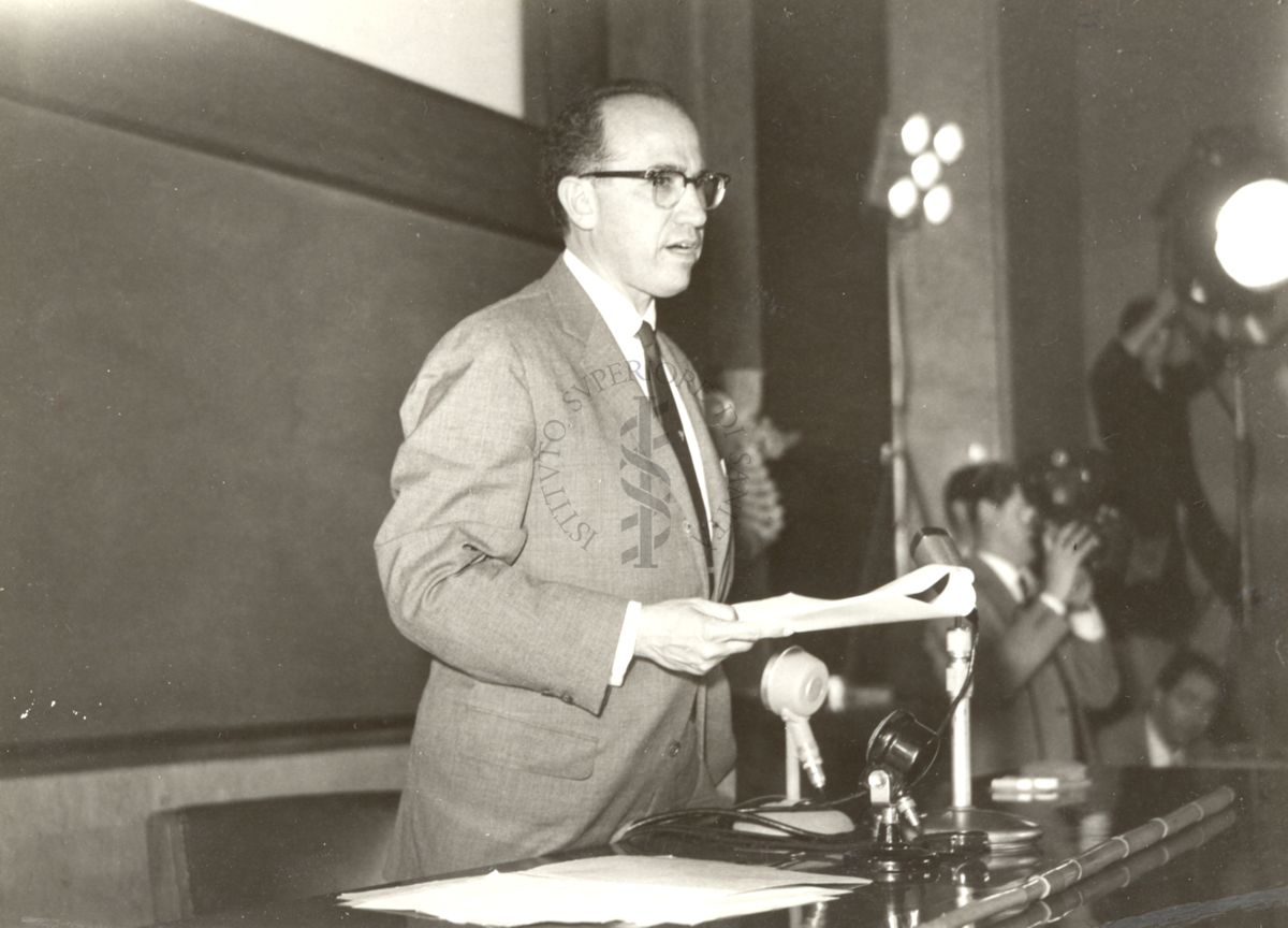 Il Prof. Salk in piedi dietro la cattedra dei relatori ripreso nell'aula magna in occasione della conferenza tenuta all'Istituto Superiore di Sanità