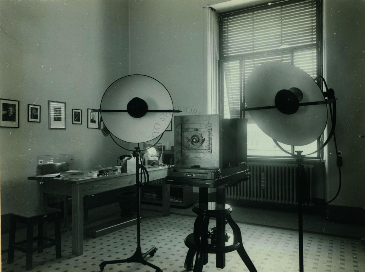 Primo piano di una parte della sala di posa del laboratorio fotografico  dove possiamo notare due lampade per la ripresa e una macchina fotografica in legno sul cavalletto