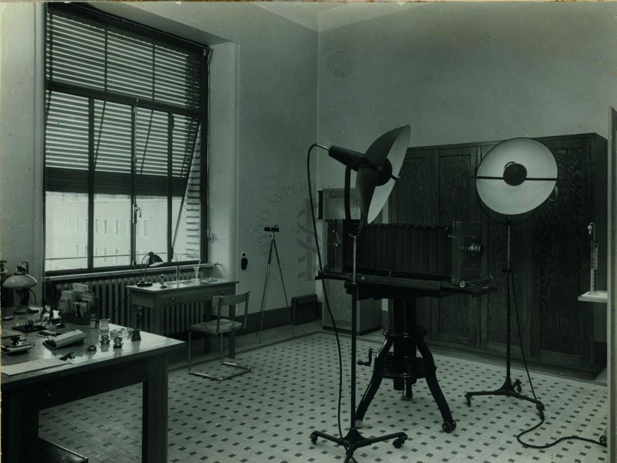 Primo piano di una parte della sala di posa del laboratorio fotografico  dove possiamo notare due lampade per la ripresa e una macchina fotografica in legno sul cavalletto