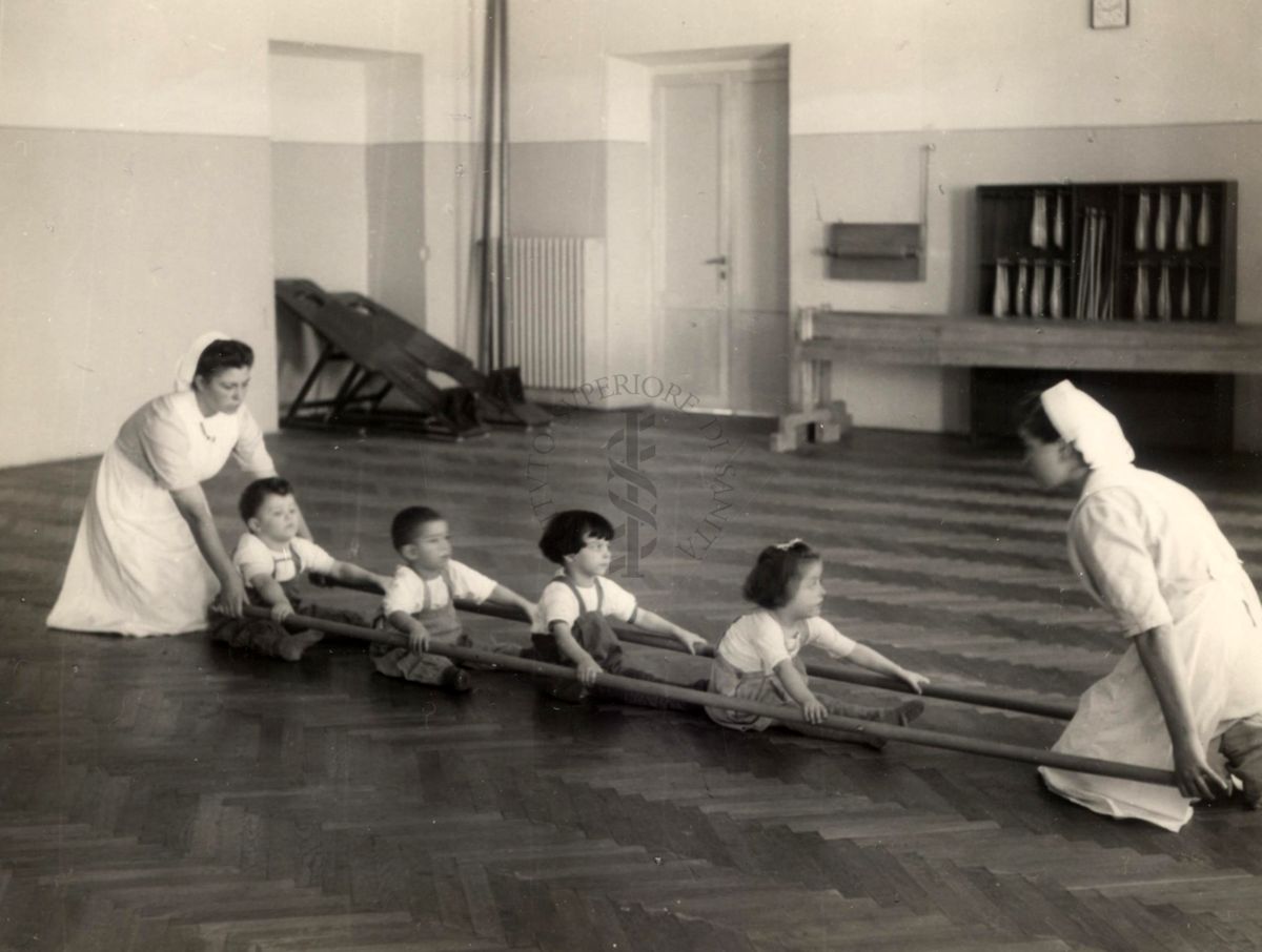 Sala per la ginnastica collettiva dell'Istituto Ortopedico "Luigi Spolverini" di Ariccia (Roma) - due infermiere sorreggono due aste per guidare i movimenti di 4 bambini