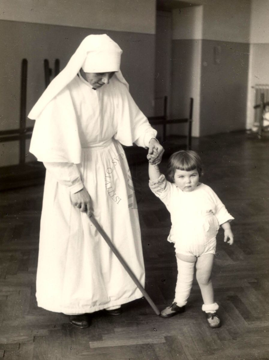 Istituto Ortopedico "Luigi Spolverini" di Ariccia - una suora infermiera tiene per mano una bambina aiutandola nella corretta postura nella sala per la deambulazione