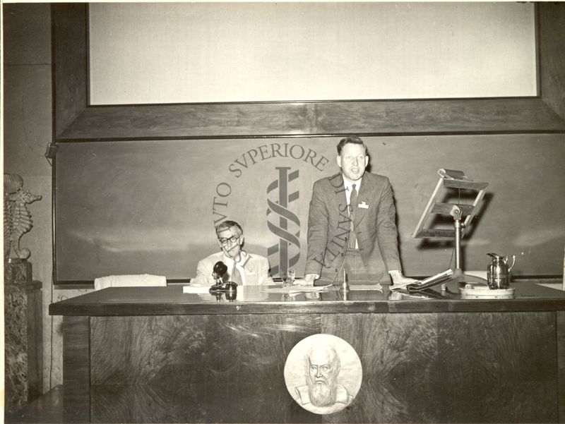 Il Prof. Frederick Charles Bawden ripreso in piedi dietro un leggio posto su un tavolo, durante la relazione tenuta al VI Congresso Internazionale di Microbiologia. Alla sua destra, seduto al tavolo, il Prof. K. N. Smith.