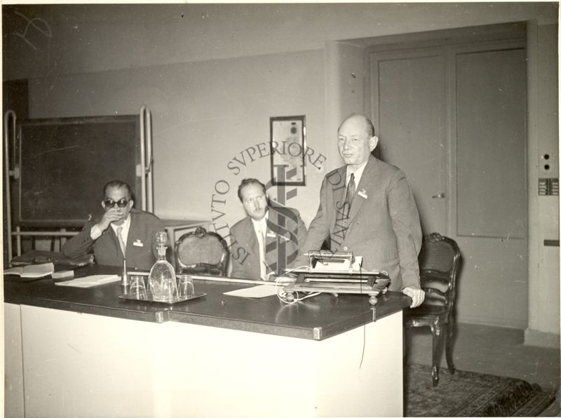 Il Prof. Henry Gerard Thornton ripreso in piedi, dietro ad un leggio posto su un tavolo, durante la relazione tenuta al VI Congresso Internazionale di Microbiologia. Alla sua destra sono seduti al tavolo altri due uomini.