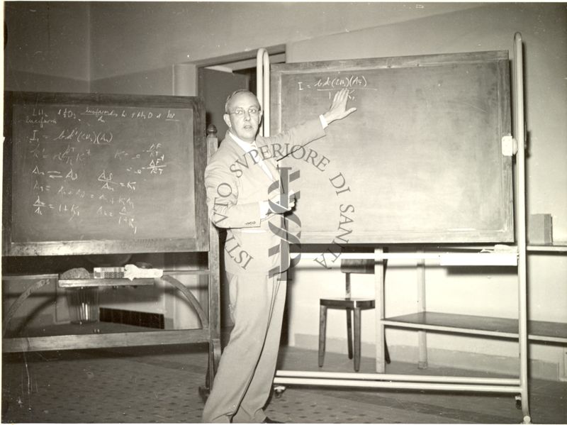 Il Prof. Frank Harris Johnson ripreso in piedi, davanti ad una lavagna mentre indica una formula matematica scrittavi sopra, durante la relazione tenuta al VI Congresso Internazionale di Microbiologia. Sullo sfondo un'altra lavagna scritta.