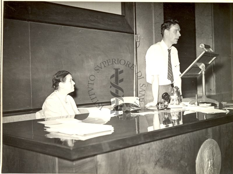 Il Prof. Robert George Everitt Murray ripreso in piedi, dietro a un leggio posto su un tavolo con le mani appoggiate al tavolo, durante la relazione tenuta al VI Congresso Internazionale di Microbiologia. Seduta al tavolo anche una donna.