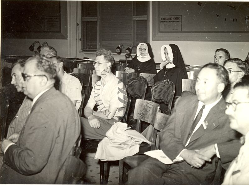 Ripresa di parte dell'uditorio seduto su sedie durante le relazioni al VI Congresso Internazionale di Microbiologia. Inquadrati: il Prof. Norman Wingate Pirie (terzo da destra in seconda fila) e Suor Marie Susanne (ultima a sinistra in ultima fila)