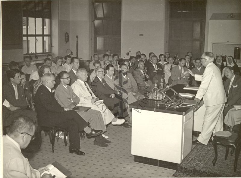 Il Prof. Selman Abraham Waksman ripreso in piedi, dietro un tavolo rivolto verso l'uditorio, durante il VI Congresso Internazionale di Microbiologia. Alla destra del Prof. Waksman ripreso un altro soggetto maschile seduto al tavolo