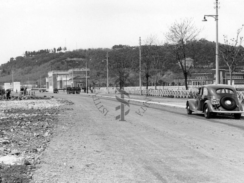 Inquadratura di una carreggiata che corre parallela al Tevere, con in fondo il Palazzo Littorio in costruzione, sulla quale transita un'autovettura targata Roma (numeri targa non visibili)