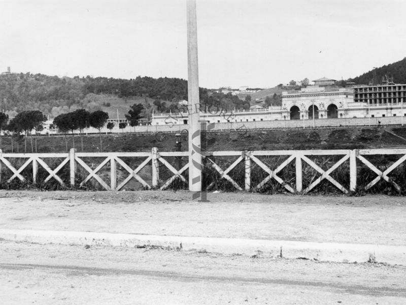 Inquadratura del marciapiede di una carreggiata che corre parallela al Tevere, con il Palazzo Littorio in costruzione sullo sfondo