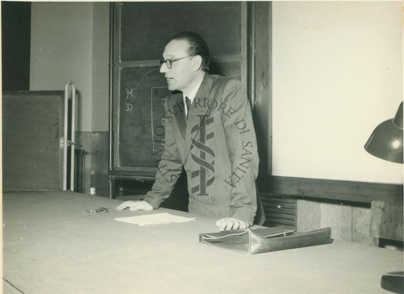 Un relatore ripreso in piedi dietro una cattedra su cui sono presenti borsa porta documenti, fogli bianchi e un orologio da polso