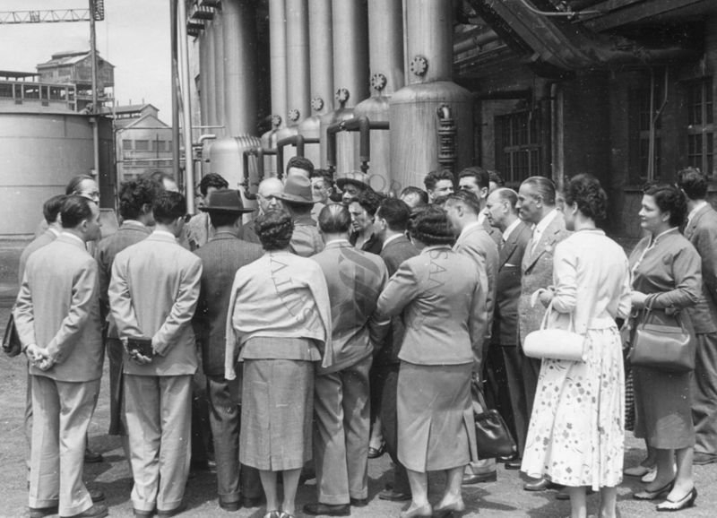 Un gruppo di uomini e donne ripresi di spalle all'esterno di una fabbrica