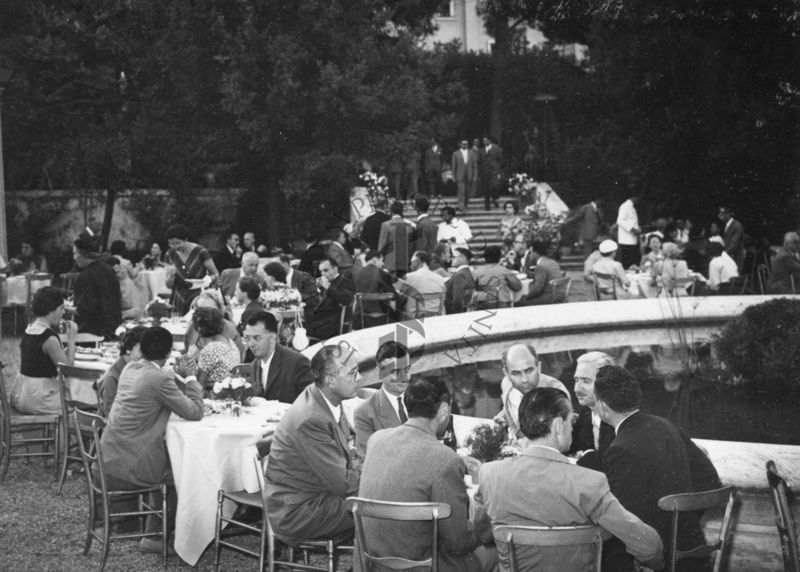 Ripresa di un lato dei tavoli allestiti in occasione del ricevimento lungo la fontana nel giardino dell'Istituto Superiore di Sanità con i partecipanti al Congresso seduti. Sullo sfondo una scalinata ed altri partecipanti in piedi.