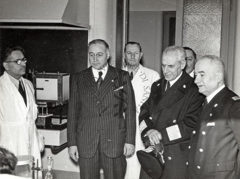 L' Ecc. Pio Calletti Sottosegretario ai  L.L.P.P. (Lavori Pubblici) ed un suo funzionario in presenza del Prof. Domenico Marotta ed alcuni ricercatori visitano un laboratorio dell' Istituto Superiore di Sanità, riconoscibili sullo sfondo: il prof. Massimo Pantaleoni e l'ingegnere capo Gregorio Birelli