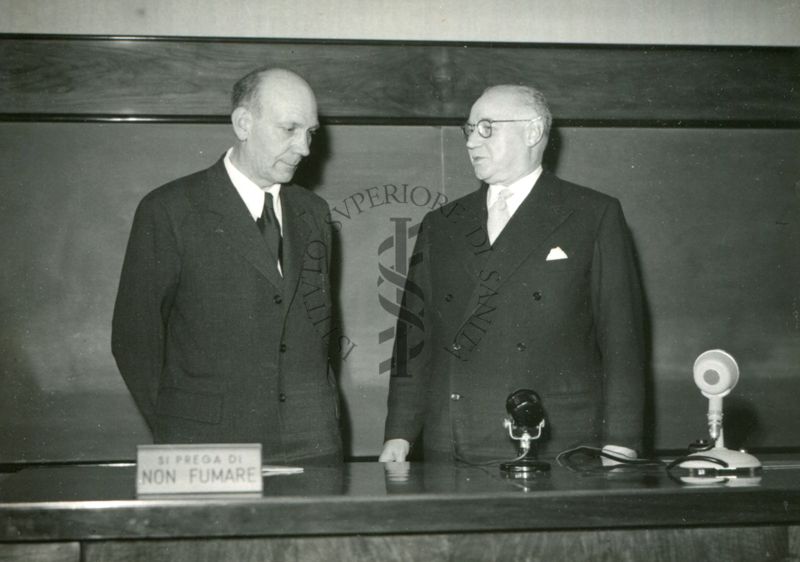 Il Prof. Holter (a sinistra) e il Prof. Bovet ripresi dietro il tavolo dei relatori durante in occasione della conferenza tenuta dal Prof. Holter nell'aula magna dell'Istituto Superiore di Sanità