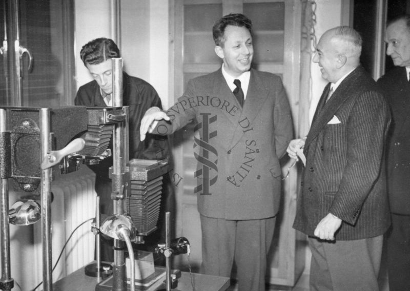 Il Ministro del Commercio con l'estero On.le Mario Martinelli (al centro della foto) in visita all'Istituto Superiore di Sanità ripreso nei laboratori fotografici dell'Istituto. Sulla sinistra un dipendente dell'Istituto in camice scuro e a destra si riconosce il Dott. Massimo Pantaleoni