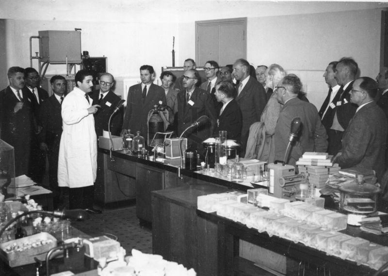 Il gruppo dei delegati UNESCO in visita all'Istituto Superiore di Sanità ripreso all'interno di uno dei laboratori, dietro ai tavoli da lavoro, intento ad ascoltare il Prof. Riccardo Milani, in camice bianco. Presente nella foto anche il Dott. Massimo Pantaleoni (dietro al gruppo all'altezza del muro bianco)