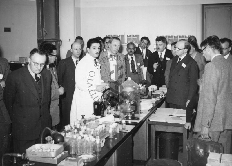 Il gruppo dei delegati dell'UNESCO in visita all'Istituto Superiore di Sanità, ripreso all'interno di un laboratorio, intorno ai tavoli da lavoro e al Prof. Milani in camice bianco.