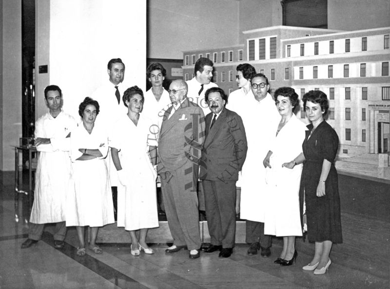 Foto di gruppo con al centro il Prof. Ernst Boris Chain e il Prof. Domenico Marotta e, primo da sinistra, in seconda fila, il Prof. Francesco Pocchiari. Sullo sfondo è visibile un plastico dell'edificio centrale di grandi dimensioni.