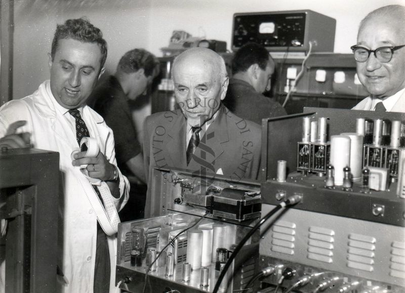 S. E. Giovanni Battista Bertone (al centro della foto) in un laboratorio dell'Istituto Superiore di Sanità in compagnia del prof. Domenico Marotta (lato destro) e il prof. Francesco Pocchiari (lato sinistro).