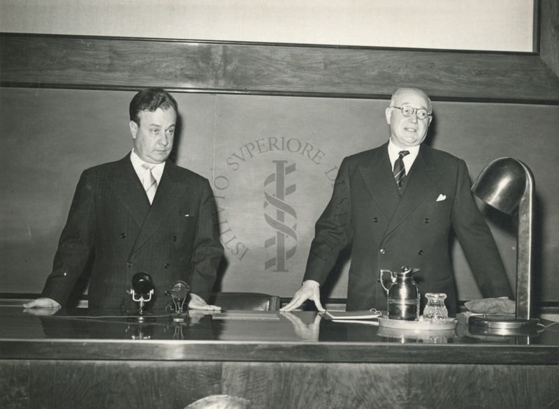 Il Prof. Carlos Chagas (a sinistra) e il Prof. Domenico Marotta (a destra) ripresi in piedi dietro al tavolo dei relatori in occasione della conferenza tenuta dal Prof. Chagas all'Istituto Superiore di Sanità