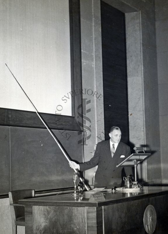 Il Prof. H. Corwin Hinshaw ripreso in piedi, dietro al tavolo dei relatori, mentre con un'asta indica la lavagna, durante la conferenza tenuta all'Istituto Superiore di Sanità.