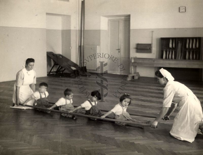 Sala per la ginnastica collettiva dell'Istituto Ortopedico "Luigi Spolverini" di Ariccia (Roma) - due infermiere sorreggono due aste per guidare i movimenti di 4 bambini