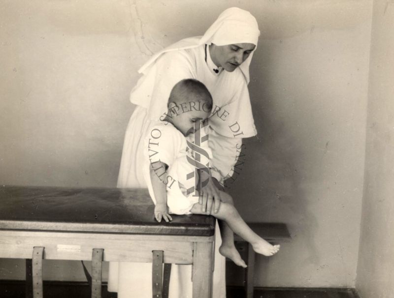 Istituto Ortopedico "Luigi Spolverini" di Ariccia - Suora-infermiera che aiuta un bambino a fare esercizi seduto su un lettino nella sala di ginnastica segmentaria