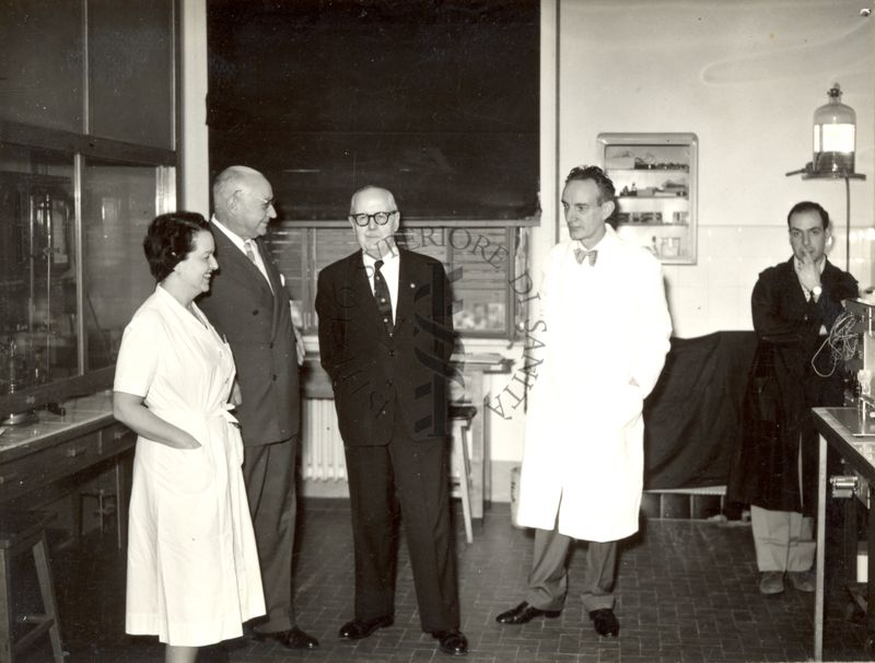 Il Prof. Bernard Houssay (vestito scuro, al centro) visita l'Istituto Superiore di Sanità, accompagnato a sinistra del Prof. Domenico Marotta e dalla Dr.ssa Filomena Nitti; mentre, a destra c'è il Prof. Daniel Bovet (camice bianco) ed un assistente di laboratorio (camice scuro)