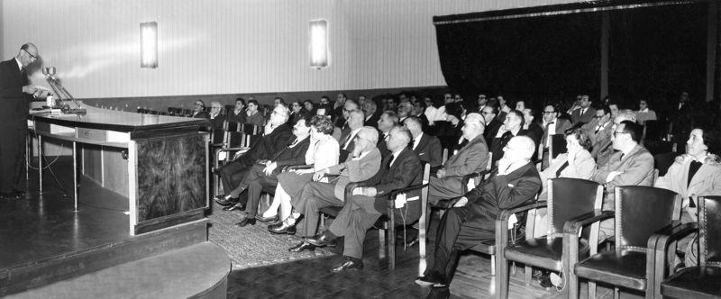 Conferenza il aula magna dell'Istituto Superiore di Sanità del Prof. Dikran Dervichian a capo dei laboratori di Biofisica dell'Istituto Pasteur di Parigi; tra il pubblico si riconoscono in prima fila: il Prof. Giordano Giacomello, il Prof. Ernst Boris Chain e la sua consorte Dr.ssa Anne Beloff Chain; in quarta fila è riconoscibile il Prof. Daniel Bovet